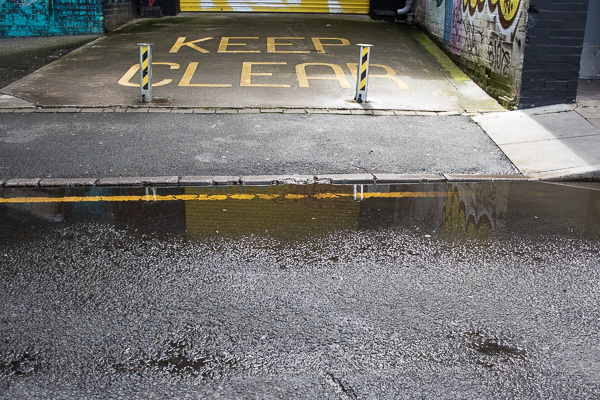Yellow writing saying Keep Clear in front of the bottom of a yellow garage door, with a puddle in front over a single yellow road line, with two yellow and black bollards and a reflection of the yellow garage door.